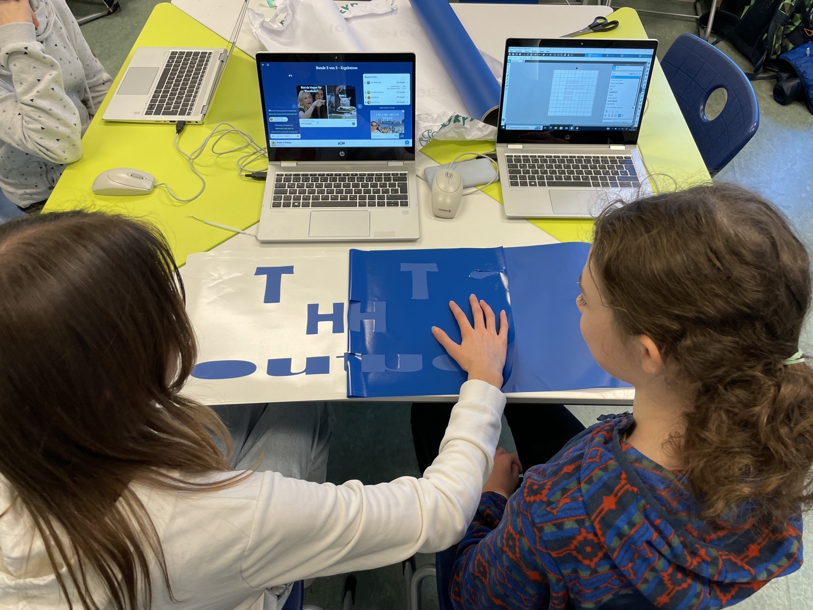 Zwei Schülerinnen schneiden an Laptops blaue Folienbuchstaben für ein Banner zu